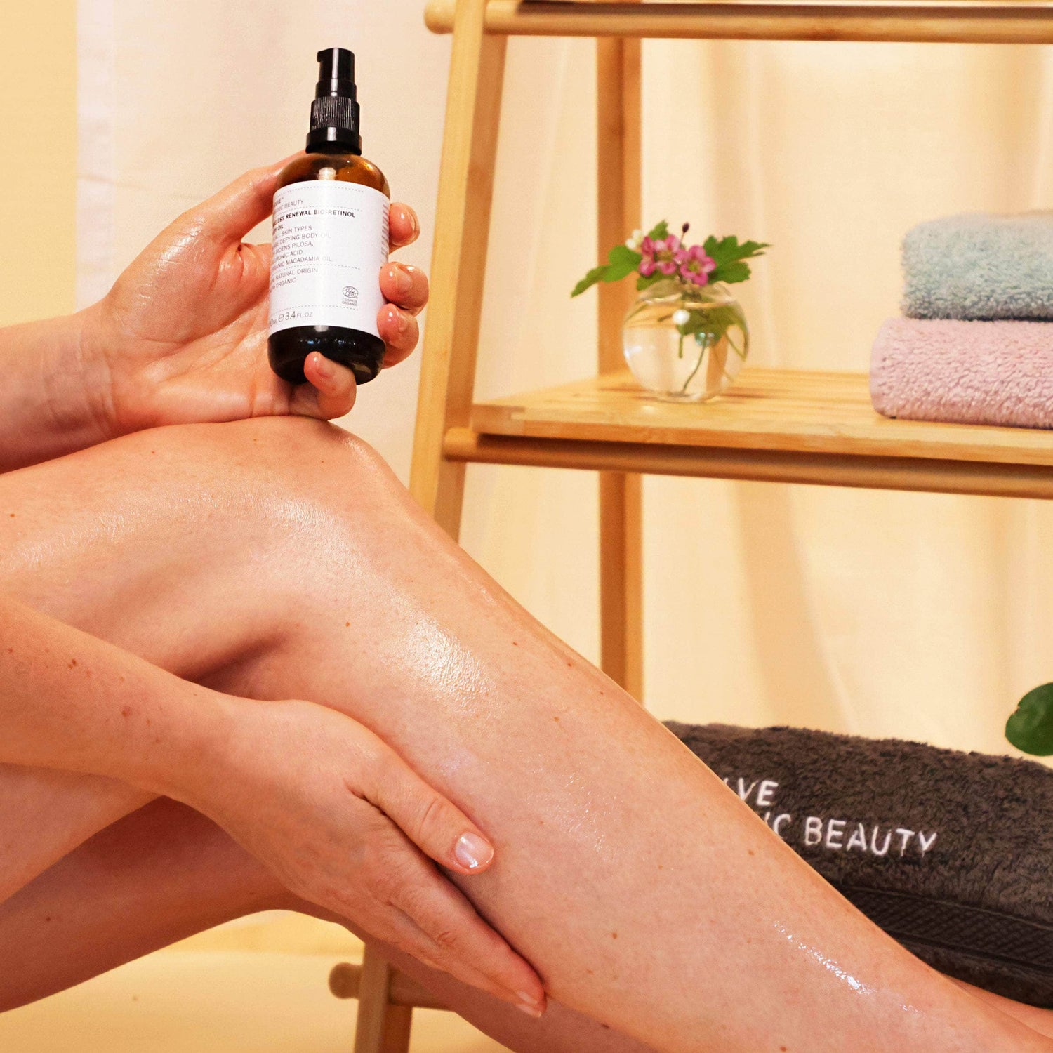 skincare model using firming body oil on leg in front of bathroom shelf with towels and flowers on it