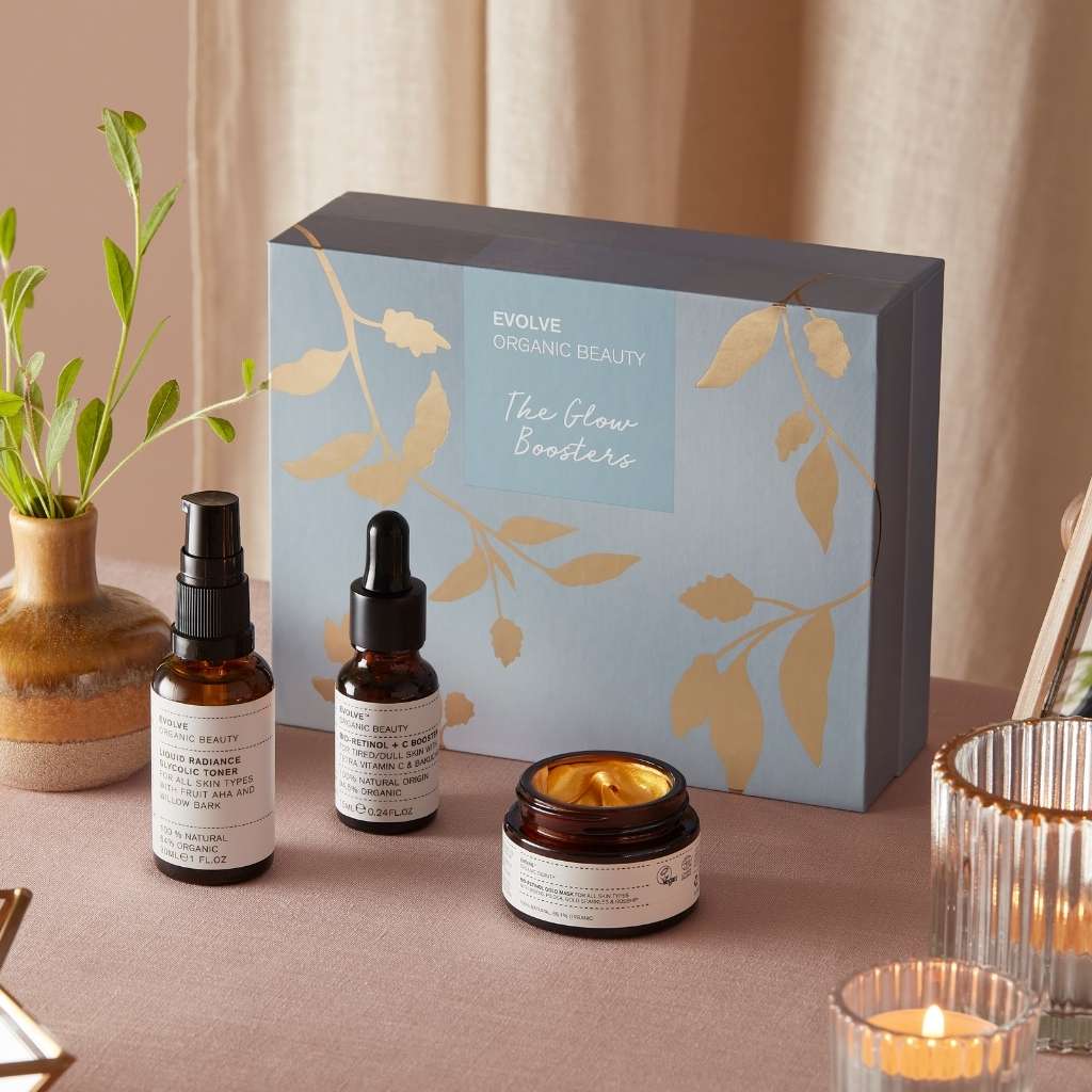 Bio-retinol gold face mask in amber glass jar, liquid radiance glycolic toner in amber glass bottle, bio-retinol + c booster vitamin c serum in amber glass bottle sat on table with plant and tea lights in front of a light blue and gold foiled gift box from Evolve Organic Beauty